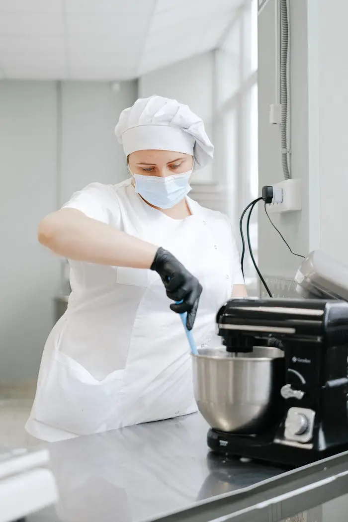 A chef wearing protective gear mixes ingredients using a stand mixer in a commercial kitchen.